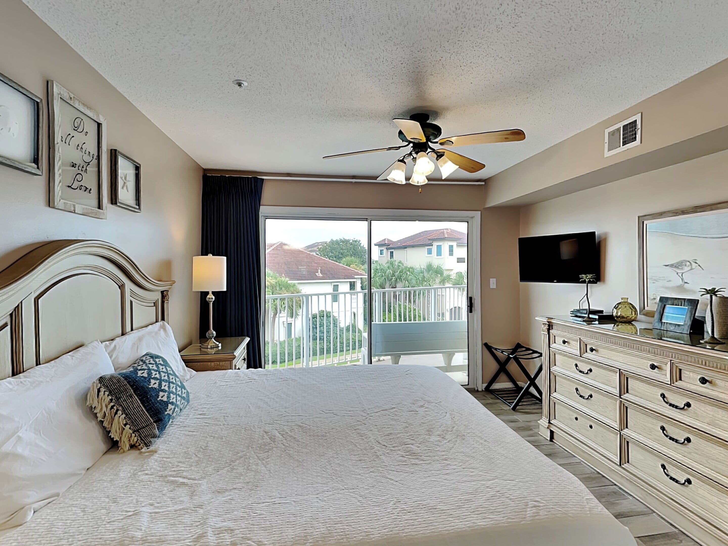 A hotel room with a bed, a ceiling fan, a TV, a dresser, and a balcony with a view of houses.
