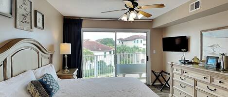 A hotel room with a bed, a ceiling fan, a TV, a dresser, and a balcony with a view of houses.