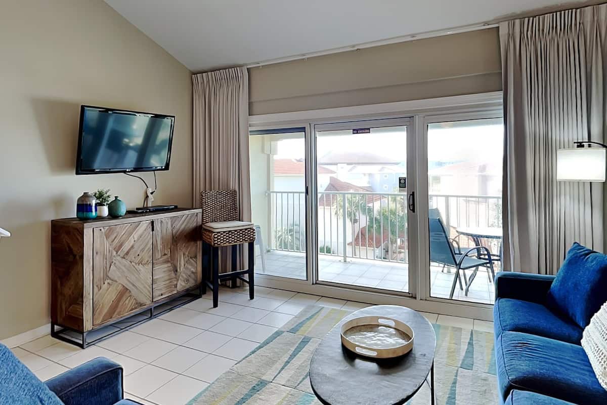 A living room with a TV, a wooden cabinet, a blue sofa, a small table, and a balcony with a view.