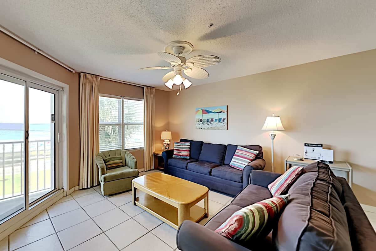 A living room with a sofa, armchair, and coffee table, a ceiling fan, and a view of the ocean through large windows.