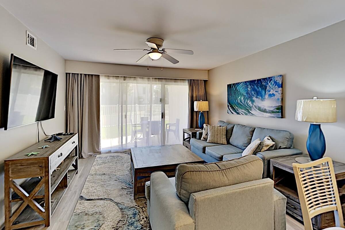 A spacious living room with a ceiling fan, a flat-screen TV, a sofa, a coffee table, and a large window with curtains.