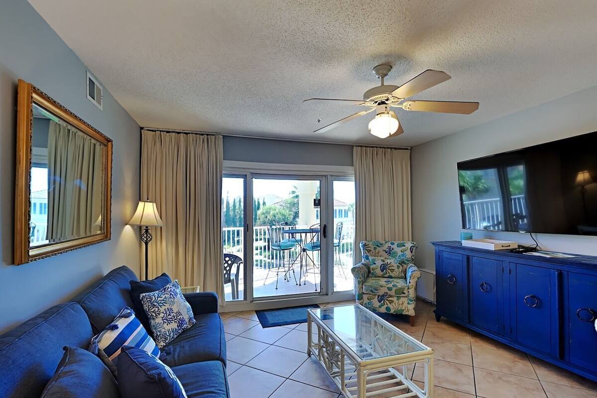 A living room with a sofa, a glass coffee table, a ceiling fan, a TV, and a view of the outside.
