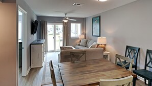 A modern living room with a wooden dining table, a sofa, and a TV mounted on the wall. - Gulf Shores Plantation II by Southern Vacation Rentals (Gulf Shores)