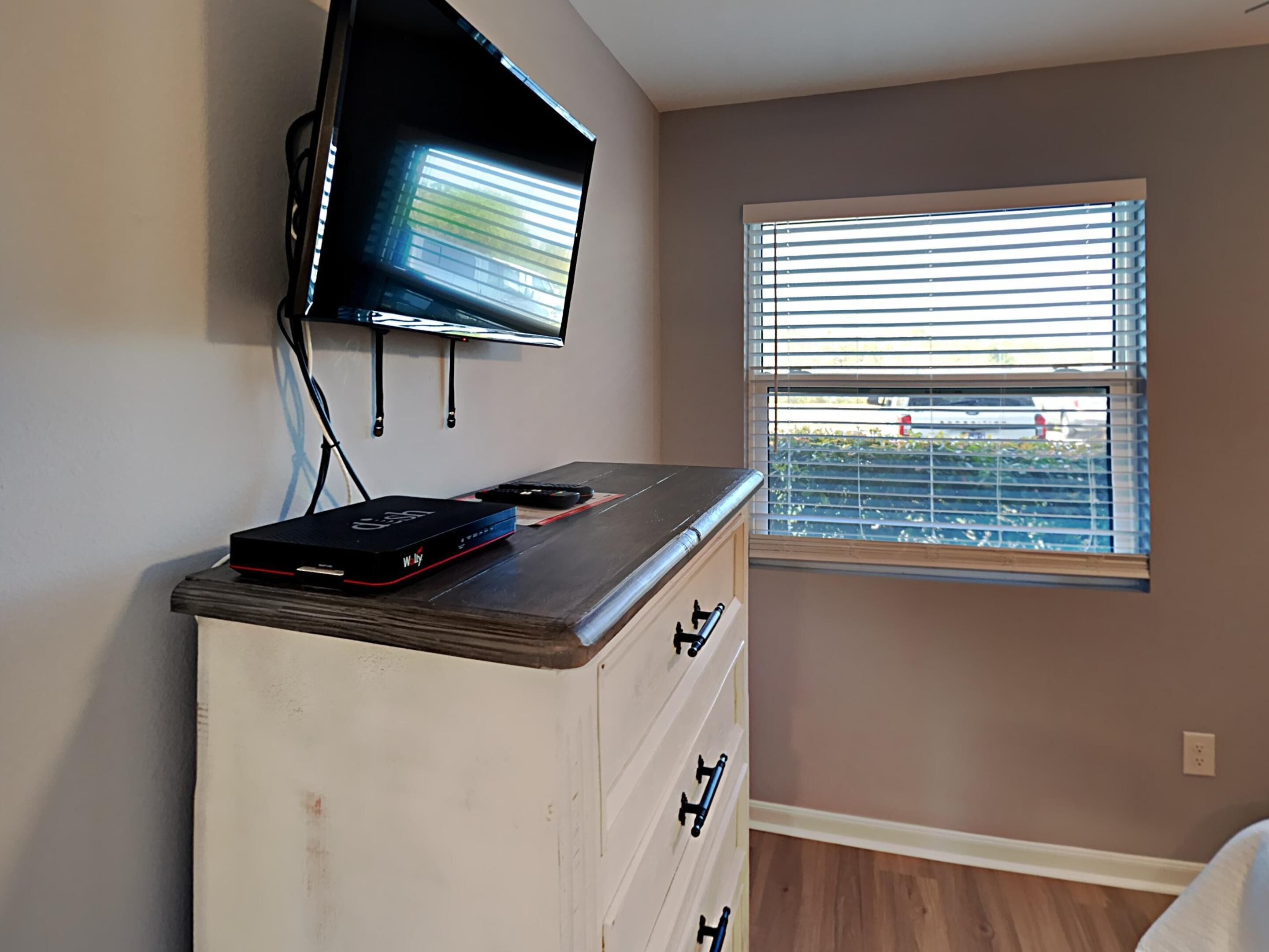 Une chambre dotée d’un téléviseur à écran plat fixé au mur, d’une commode en bois avec des tiroirs et d’une fenêtre équipée de stores.