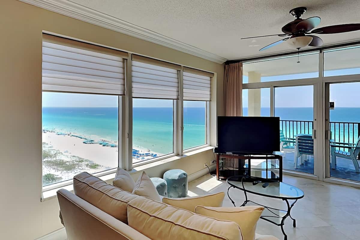 A living room with a large window overlooking a beach, a ceiling fan, and a flat-screen TV on a coffee table.