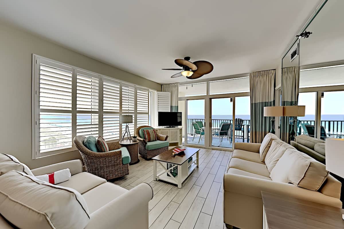 A spacious living room with a large sofa, a smaller sofa, a coffee table, and a television. There are wicker chairs and a balcony with a view of the ocean.