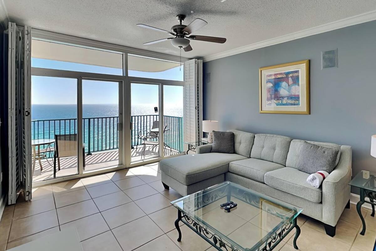 A living room with a glass coffee table, a grey sofa, a ceiling fan, and a balcony with a view of the ocean.