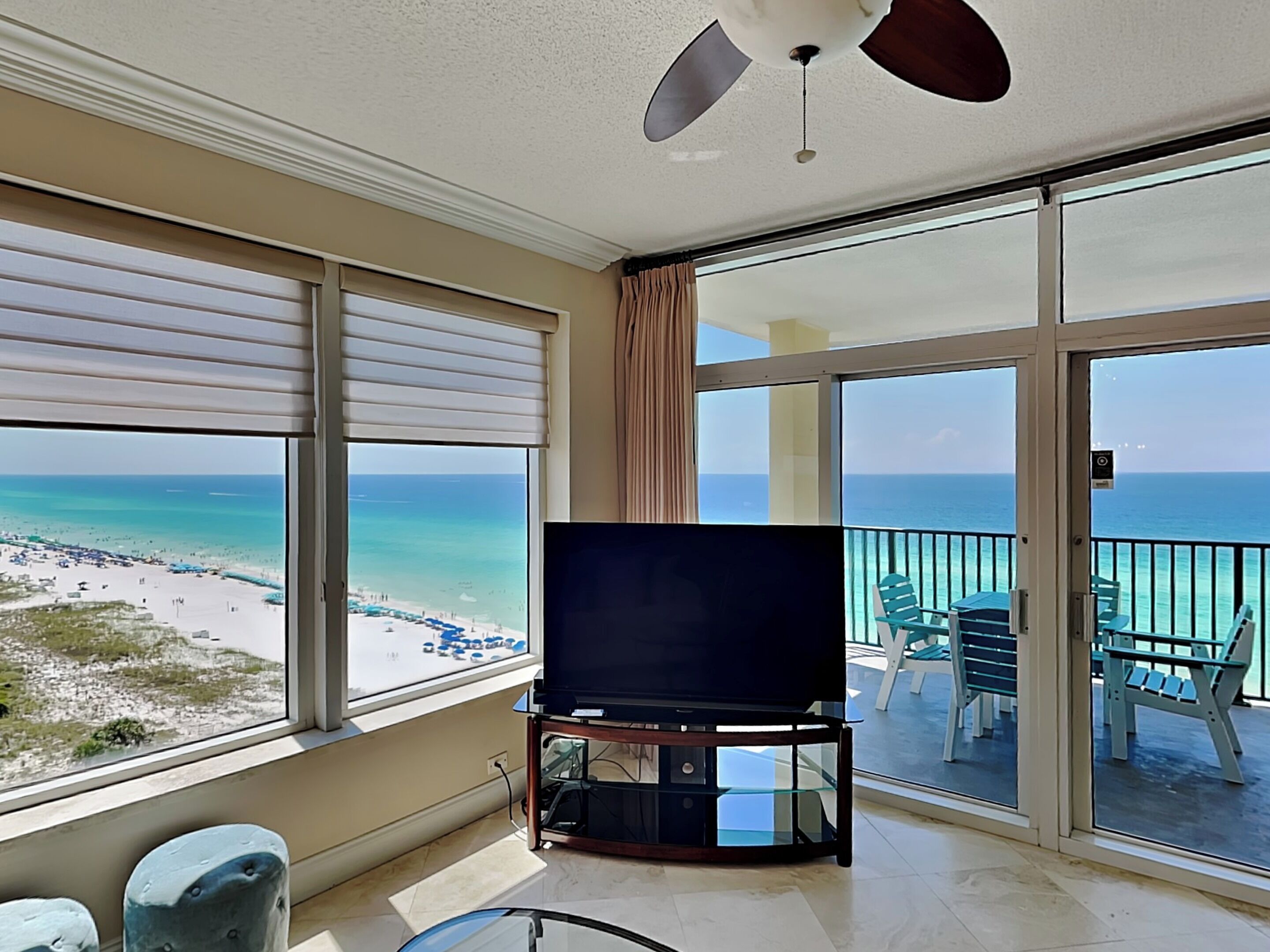 A balcony with a TV, large windows with beach views, and a ceiling fan.