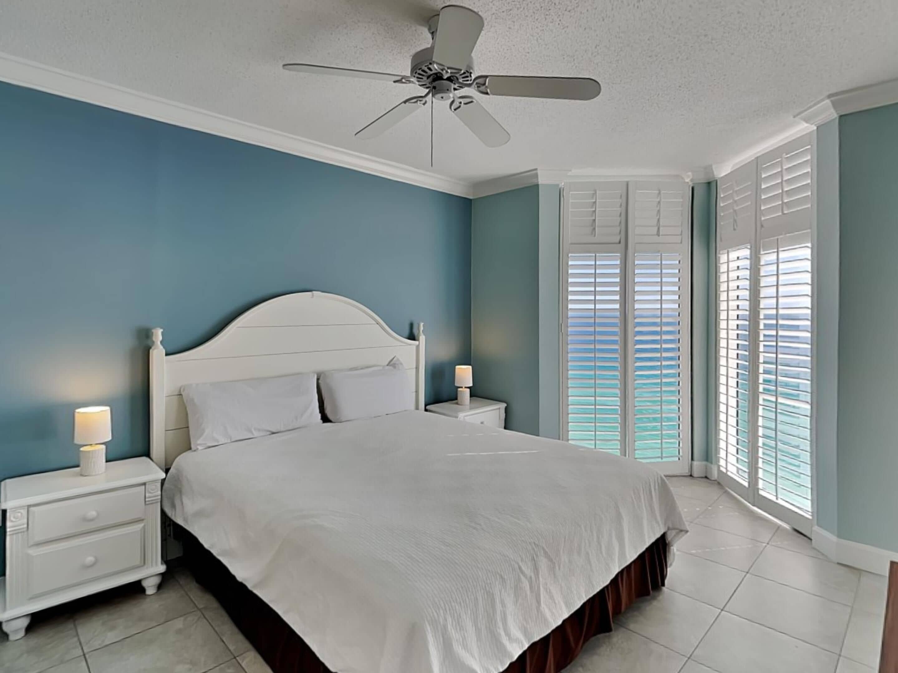 A bedroom with a white bed, two bedside tables with lamps, a ceiling fan, and a view of the ocean through the window.