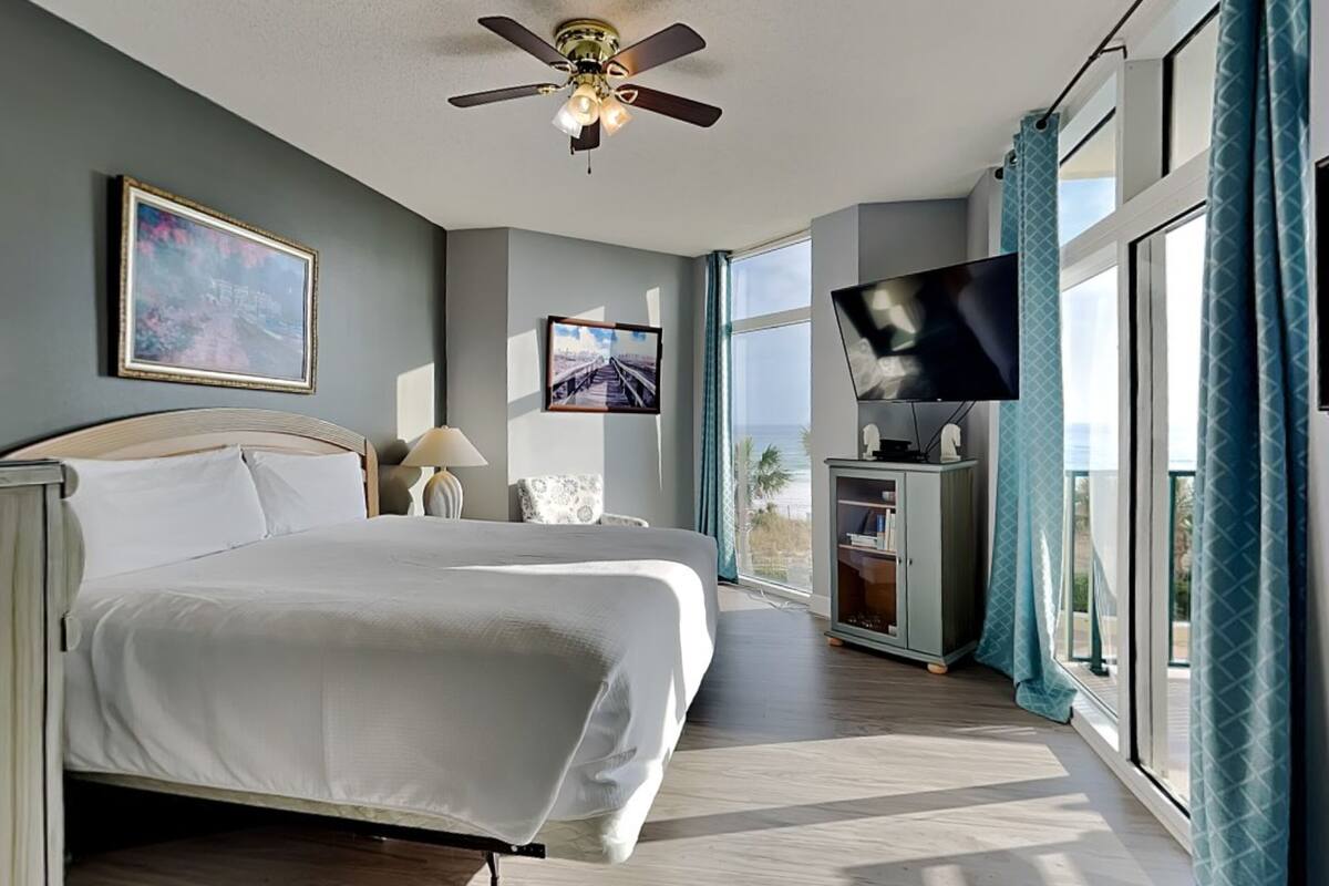 A spacious bedroom with a large bed, a ceiling fan, a TV, and a view of the ocean.