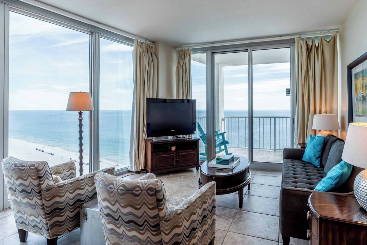 A spacious living room with a large TV, comfortable seating, and a balcony with a view of the ocean.