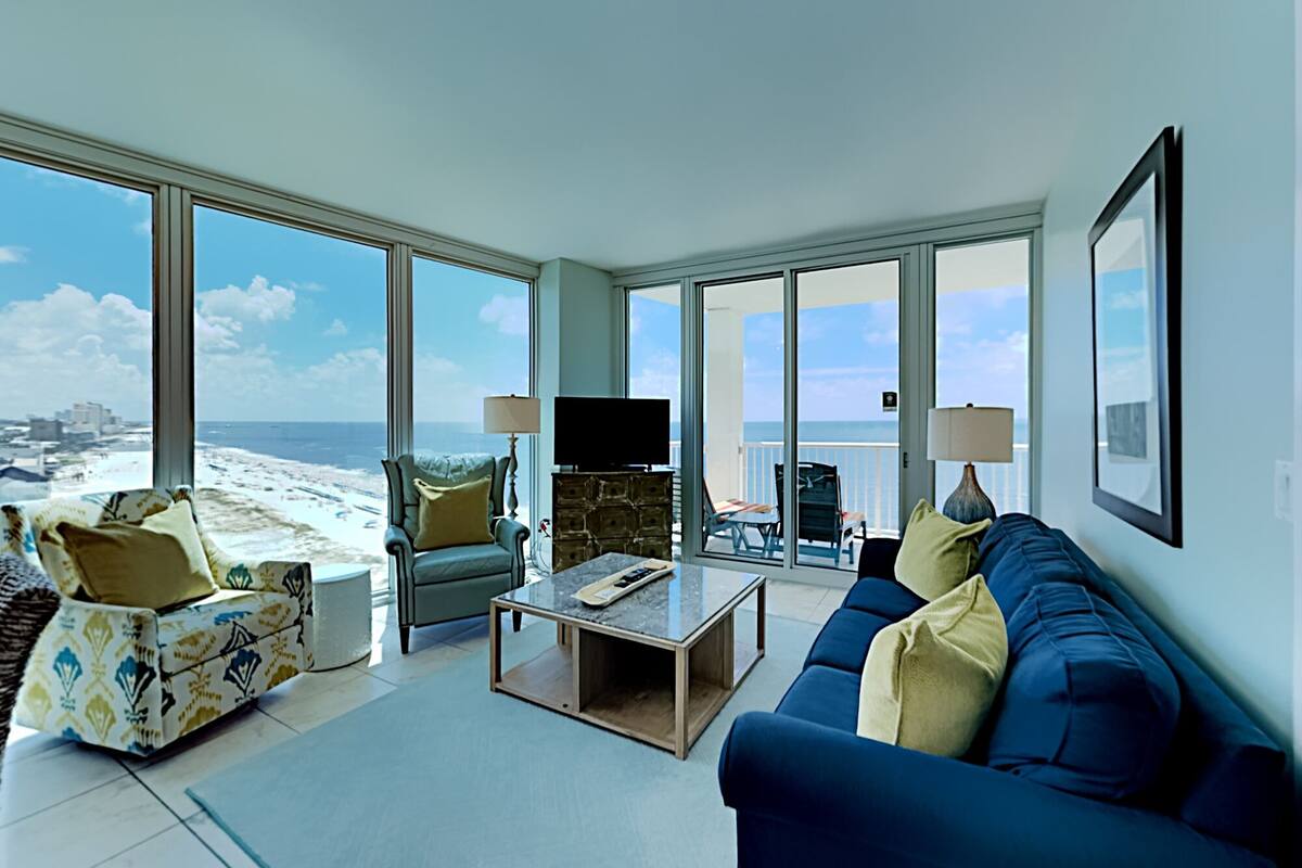 A spacious living room with a large window overlooking a beach, a flat-screen TV, and a comfortable seating area with a sofa and armchairs.