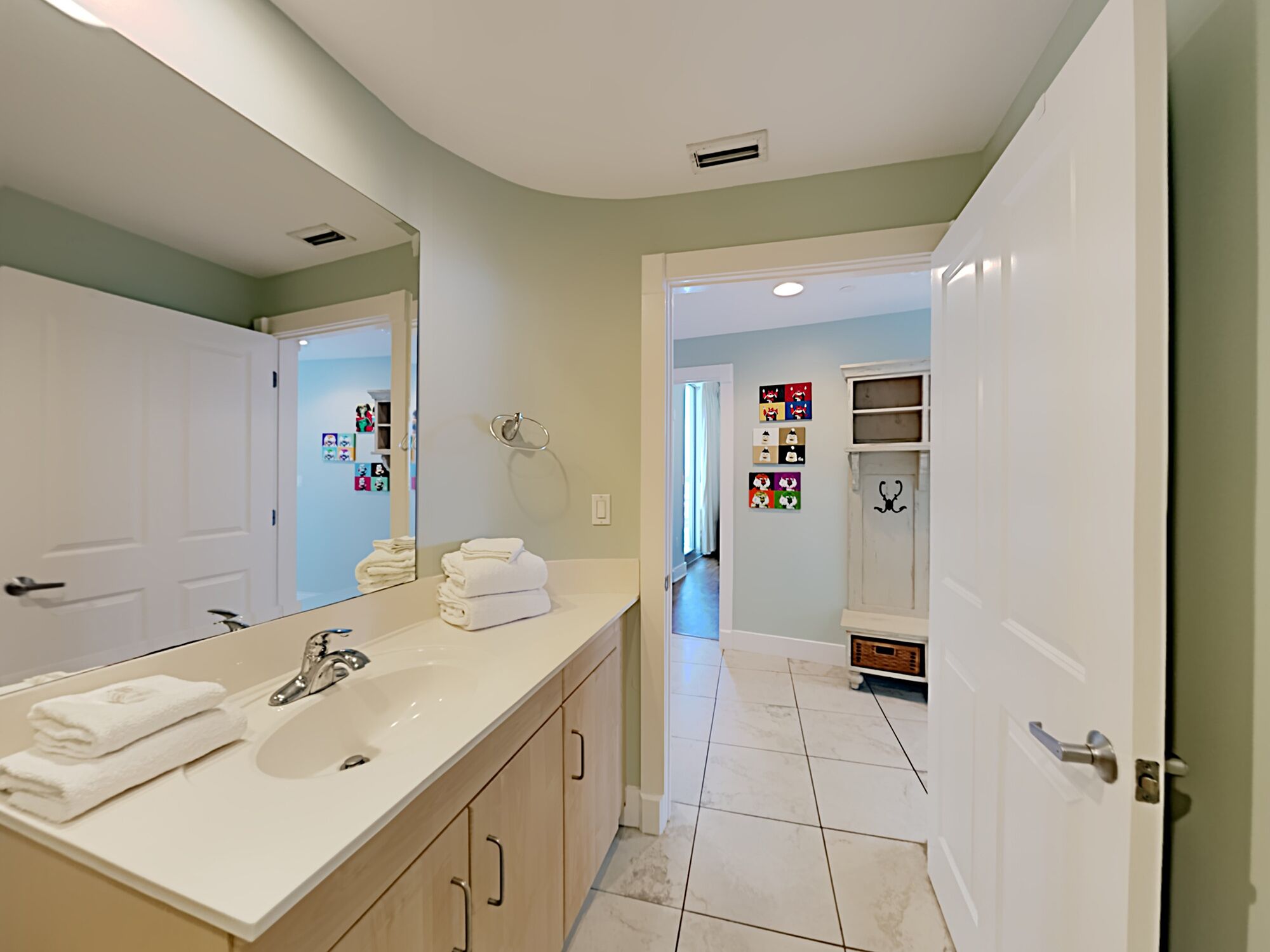 A modern bathroom with a large mirror, white sink, and wooden cabinets. There are towels stacked on the counter and a shower area visible through the door.