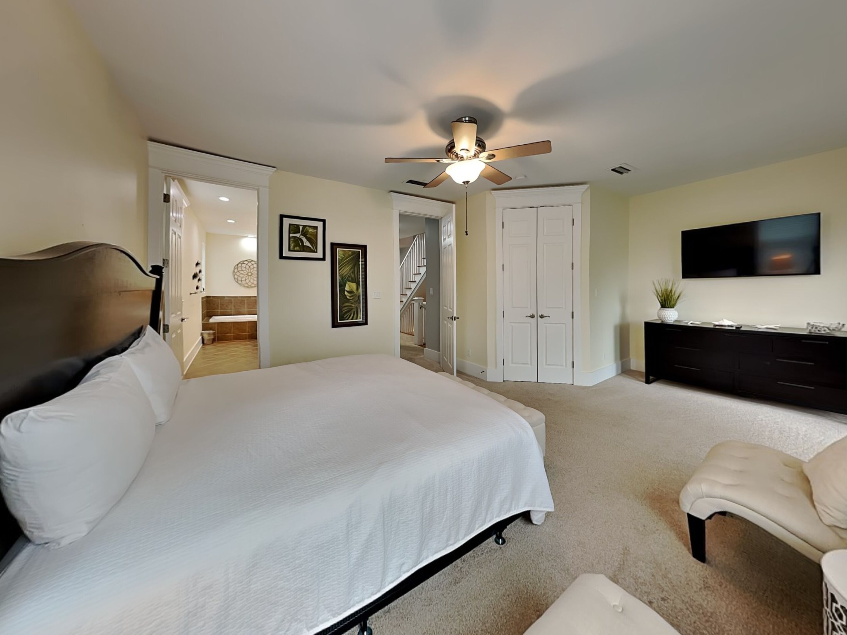 A bedroom with a bed, a ceiling fan, a TV, a dresser, and a door leading to another room.