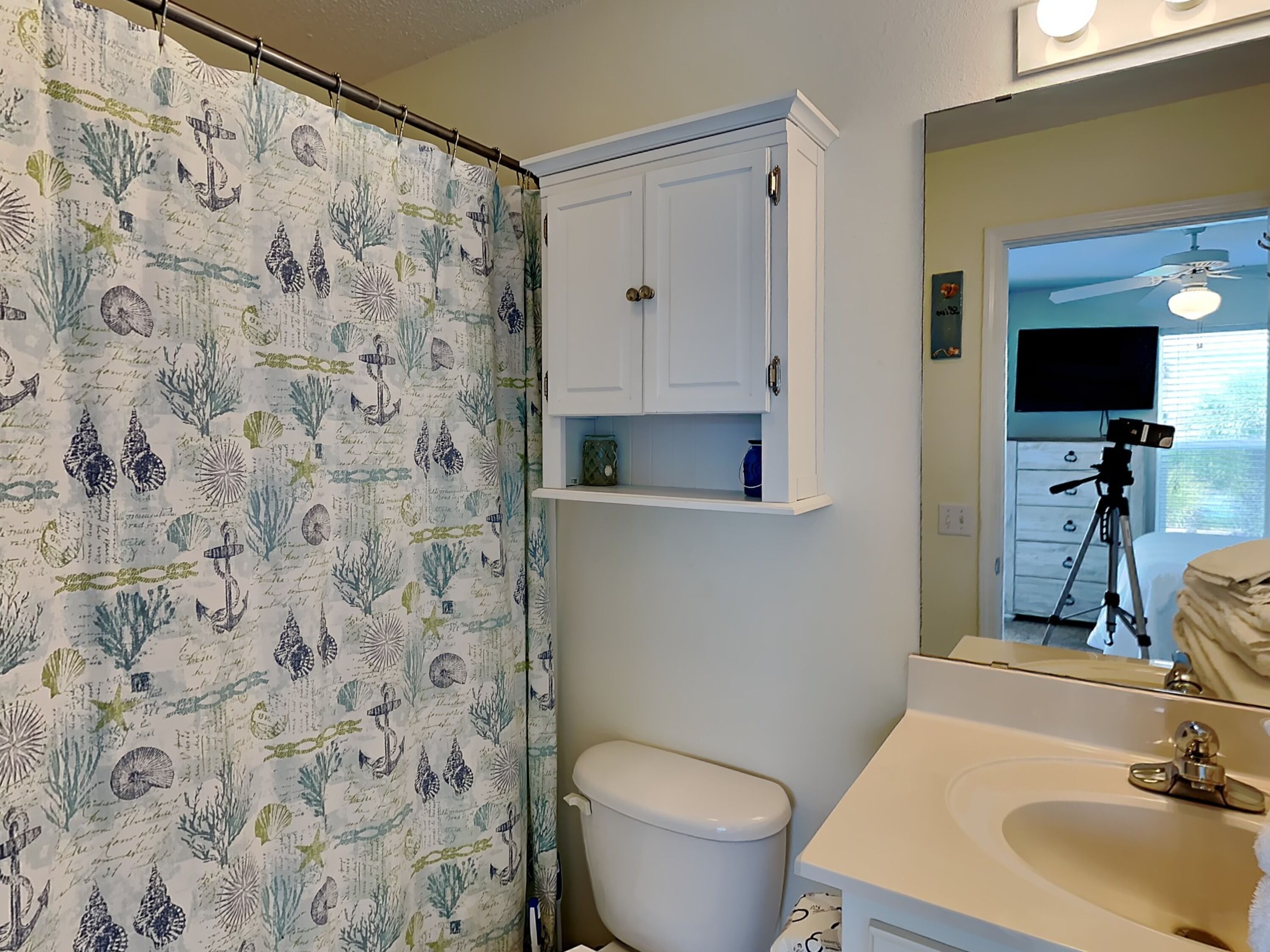 Une salle de bain avec un rideau de douche orné de motifs représentant la vie marine, des toilettes blanches, un lavabo avec un miroir et un meuble de rangement au-dessus des toilettes.