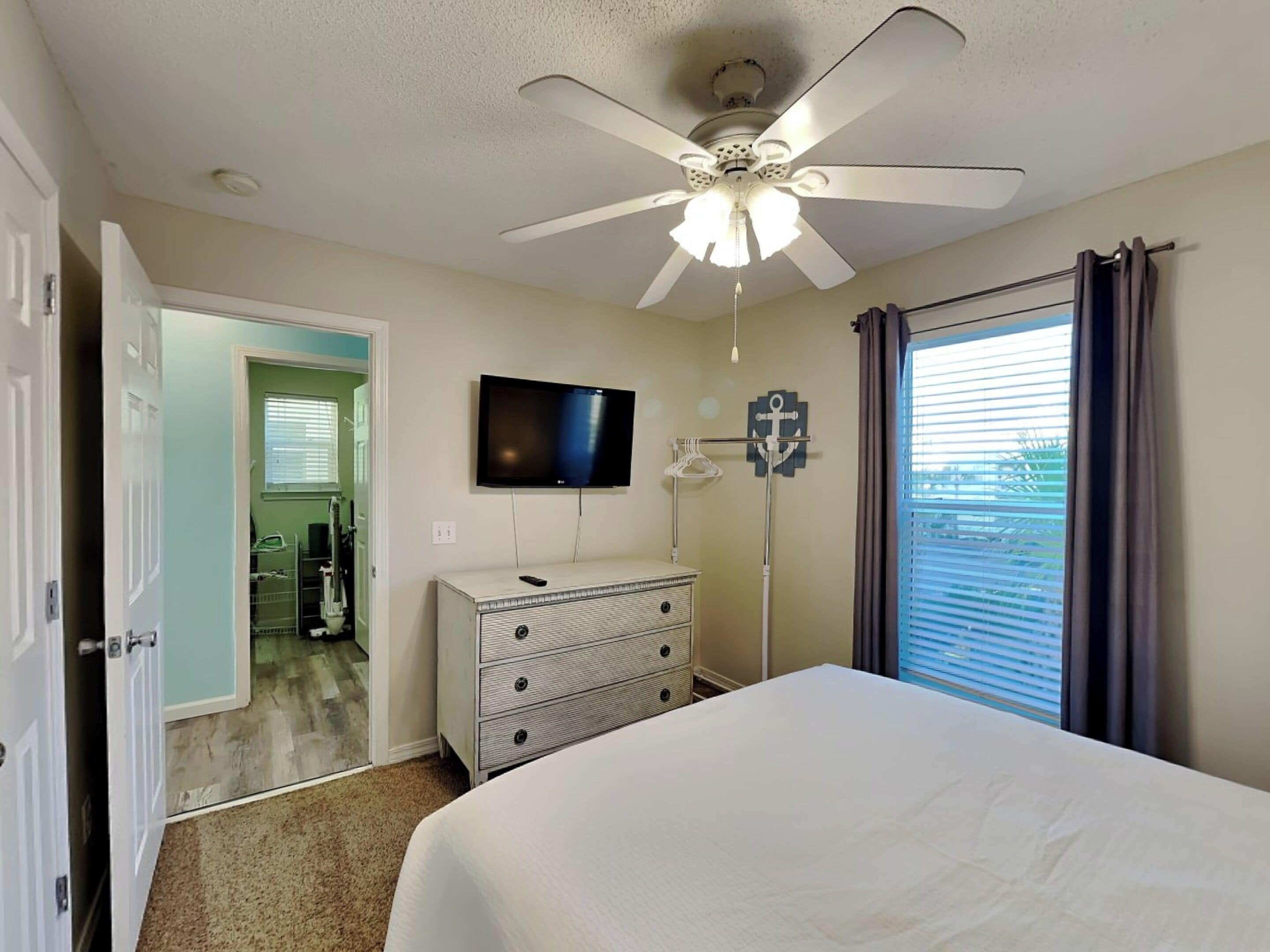 A bedroom with a bed, a ceiling fan, a TV, a dresser, and a window with curtains.