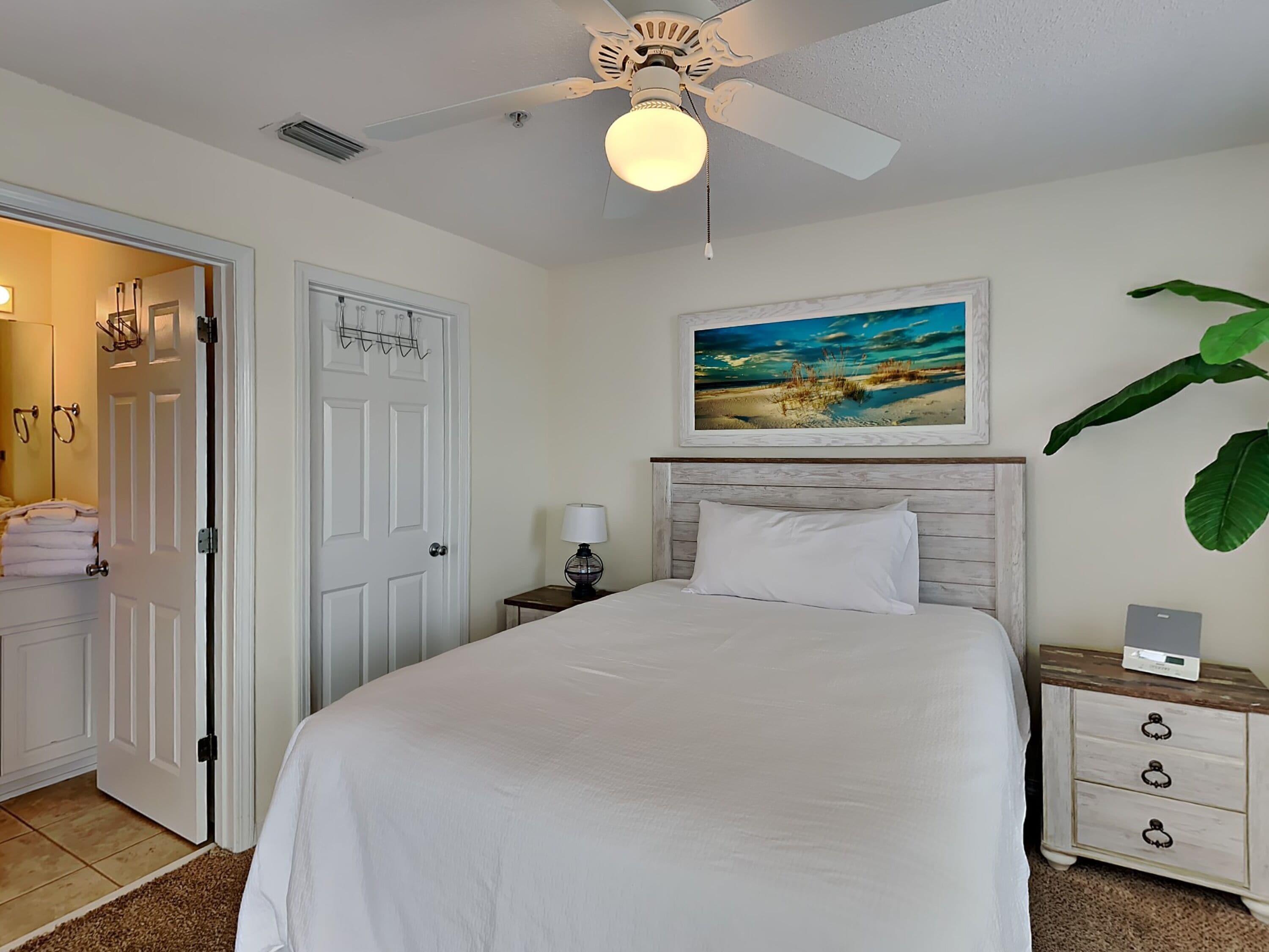 A bedroom with a bed, a nightstand, a lamp, a ceiling fan, a door, and a painting on the wall.