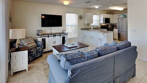 A living room with a sofa, a coffee table, and a TV. There is a kitchen area with a microwave, refrigerator, and a dining table with stools. - Gulf Island by Southern Vacation Rentals (Navarre)