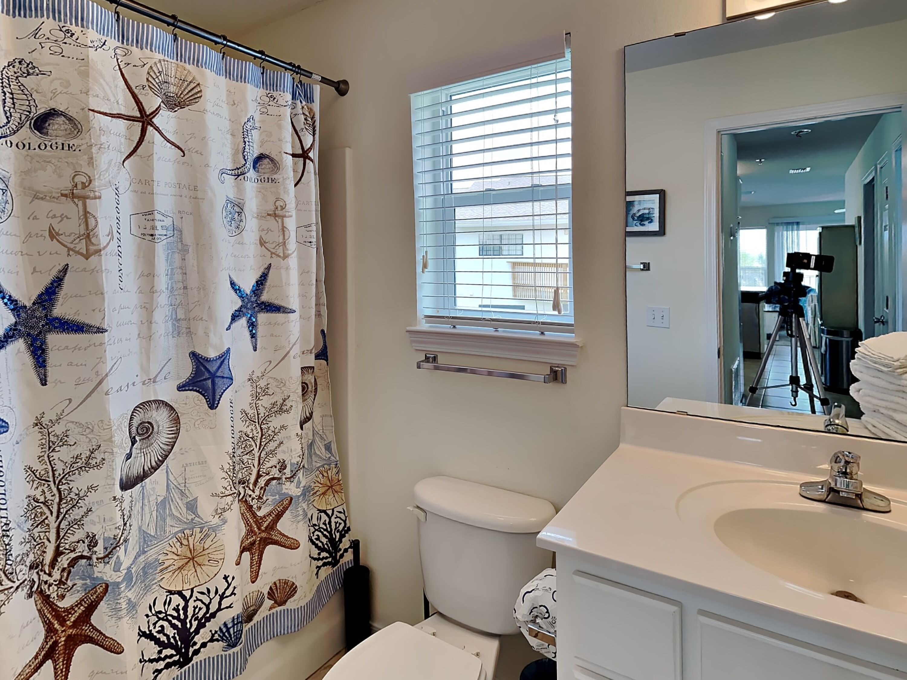 A bathroom with a nautical-themed shower curtain, a window with blinds, a toilet, and a sink with a mirror.