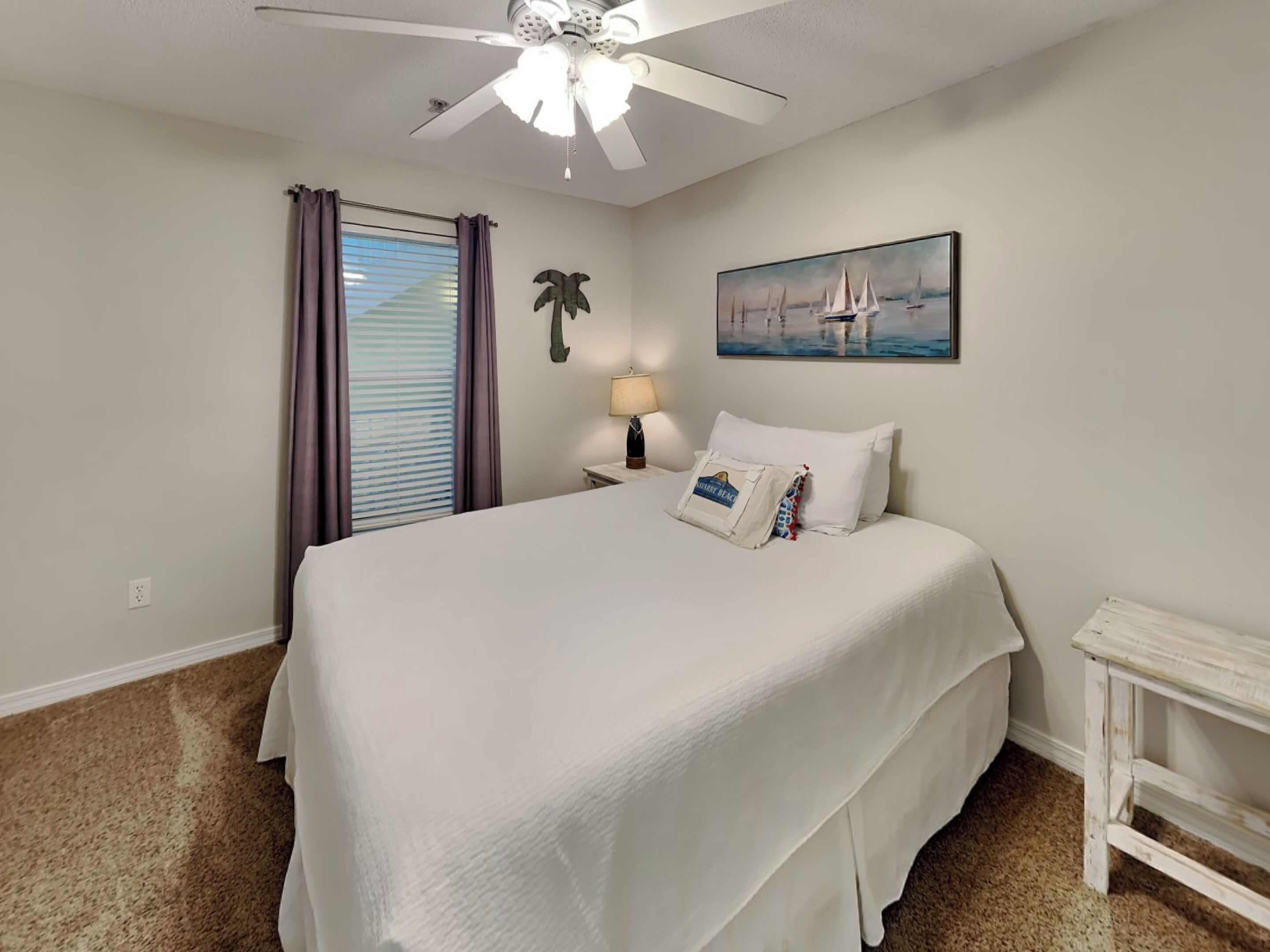 A hotel room with a large bed, a ceiling fan, a painting of sailboats, a nightstand with a lamp, and a small table.