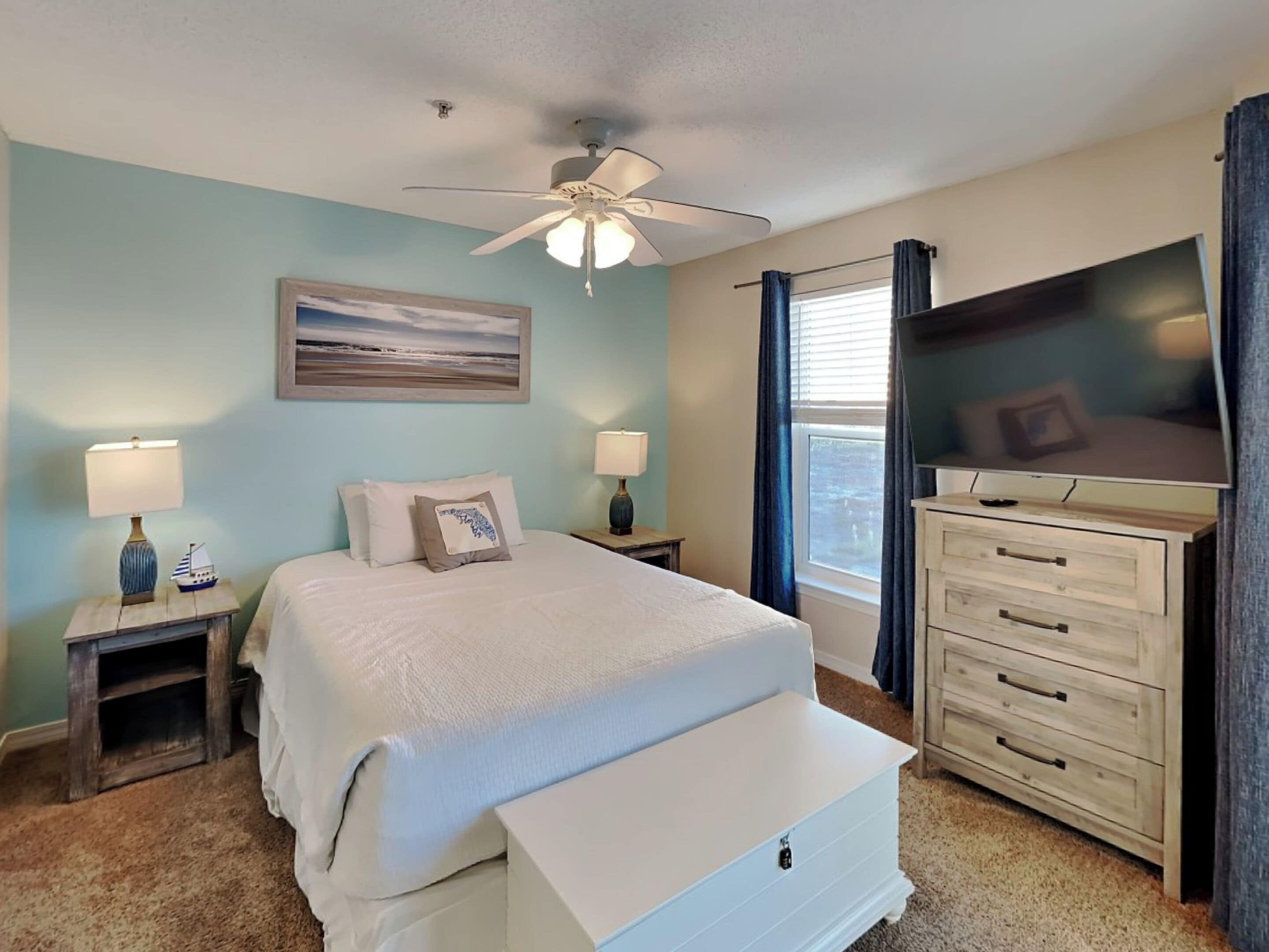 Une chambre à coucher comprenant un lit, deux tables de chevet, un ventilateur de plafond, une télévision, une commode et une fenêtre avec des rideaux.