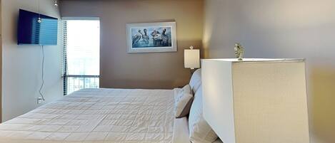 Habitación de hotel con cama, ventilador de techo, ventana con persianas, televisor montado en la pared y un cuadro.