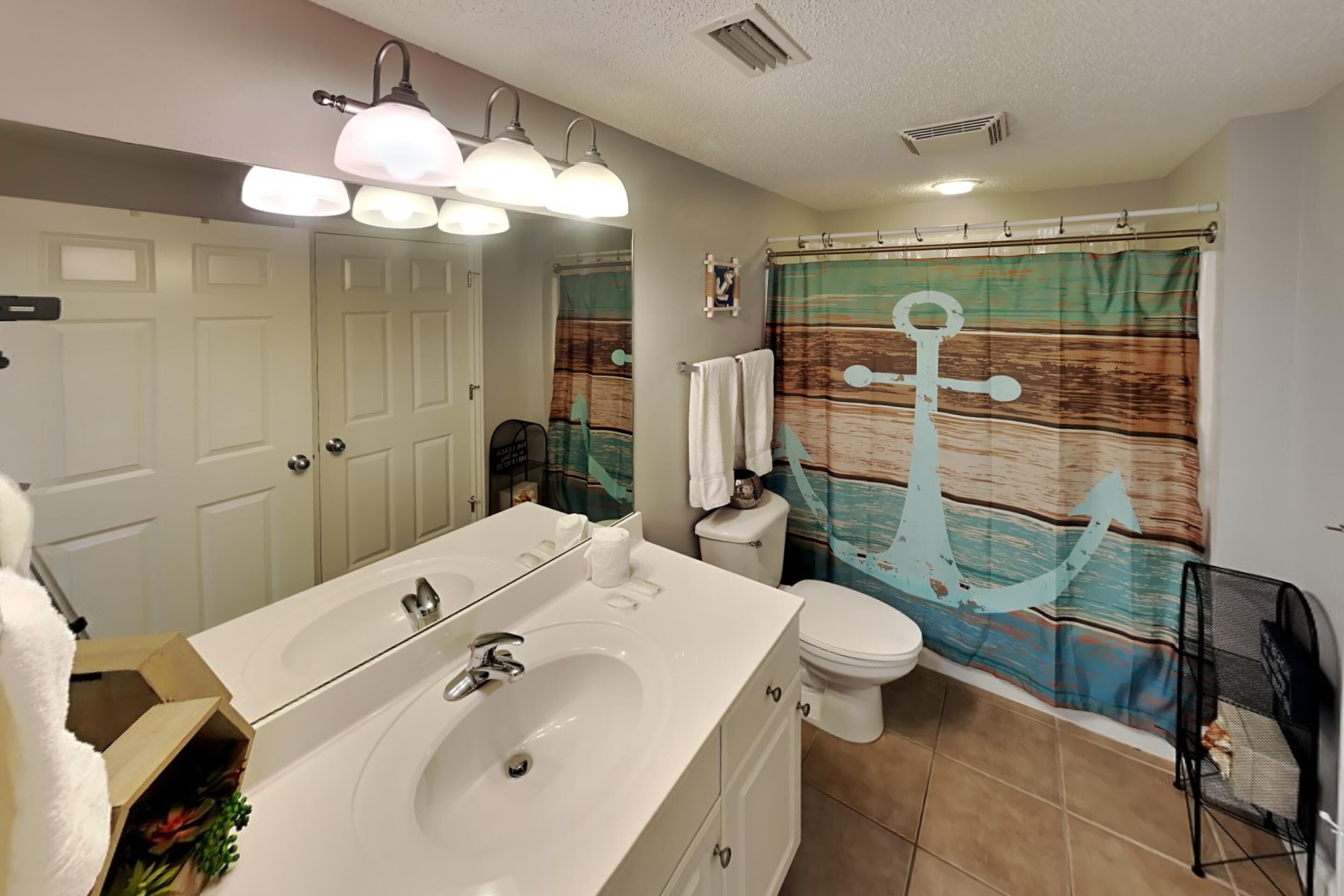 A bathroom with a white sink, two overhead lights, a shower curtain with an anchor design, and a toilet.