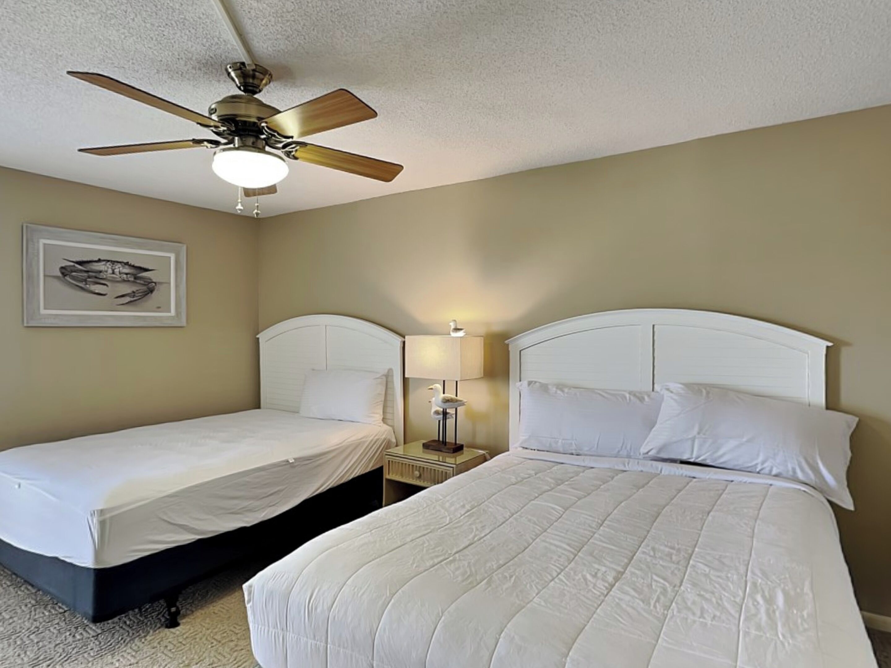 A hotel room with two single beds, a ceiling fan, a nightstand with a lamp, and a framed picture on the wall.