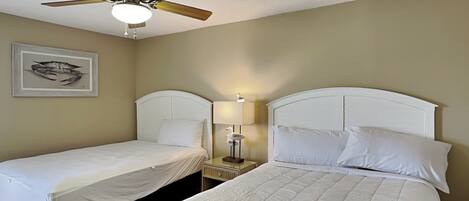 A hotel room with two single beds, a ceiling fan, a nightstand with a lamp, and a framed picture on the wall.