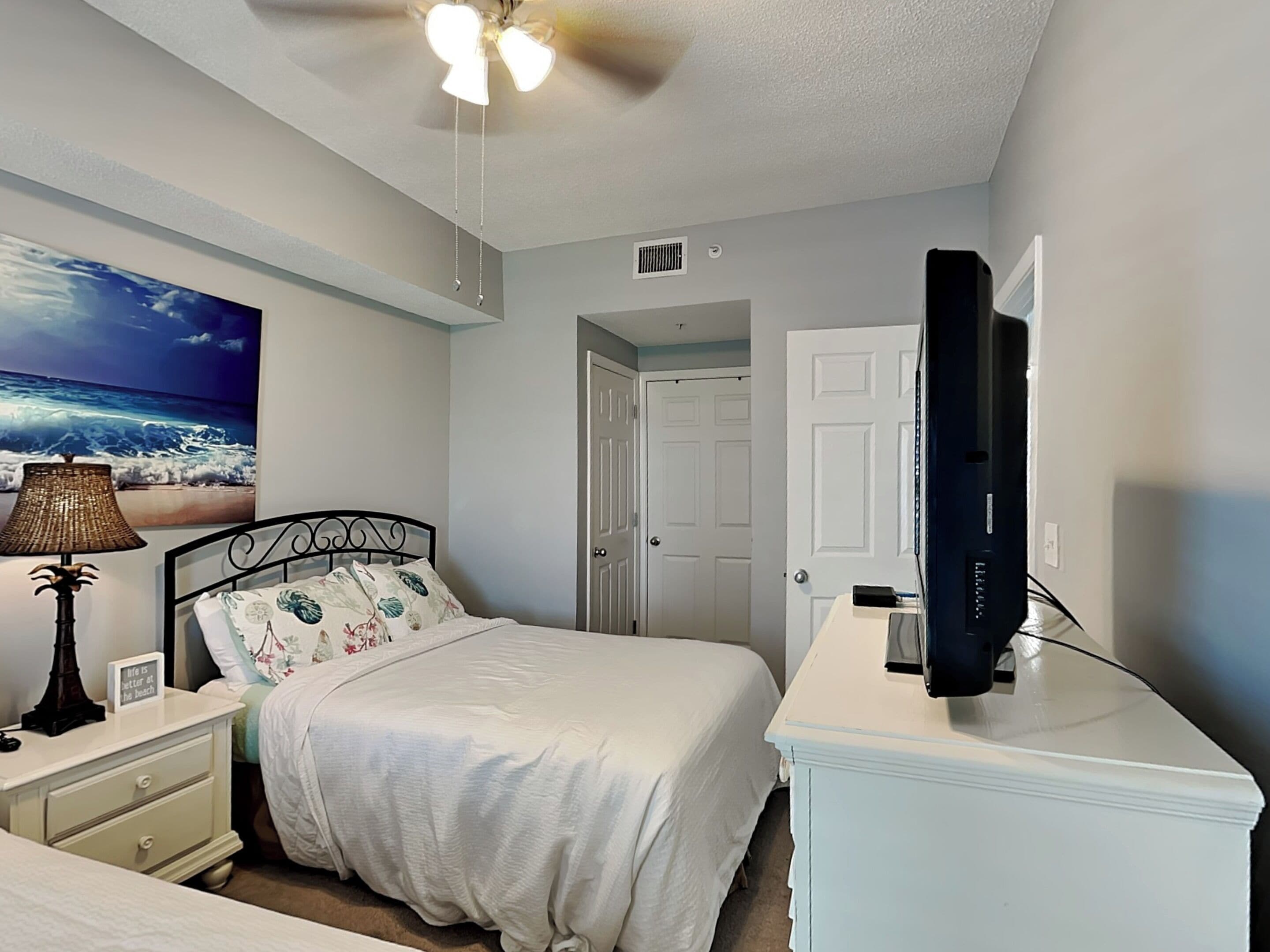 Une chambre à coucher comprenant un lit, une table de chevet, une lampe, un ventilateur de plafond, un téléviseur et une porte.