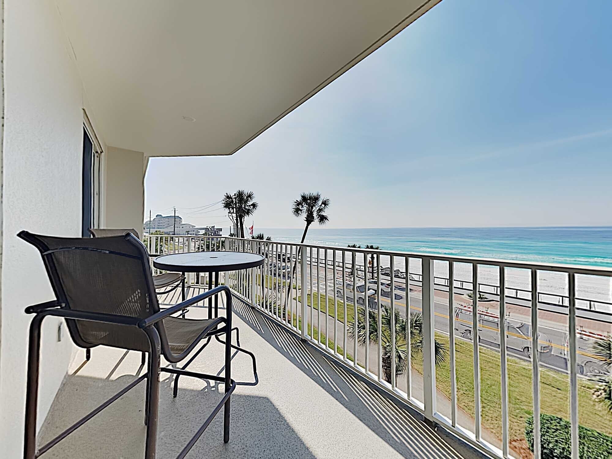 Un balcon avec une table et des chaises donnant sur une plage, offrant une vue dégagée sur l’océan et un espace de stationnement.