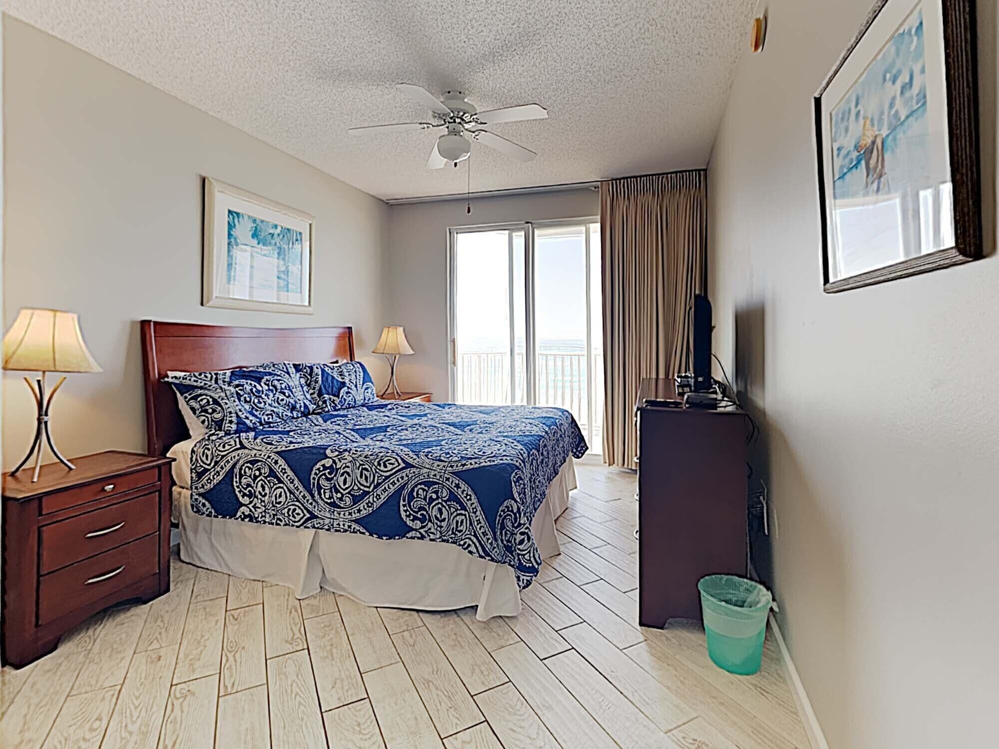 A hotel room with a bed, a nightstand, a ceiling fan, a TV, and a balcony with curtains.