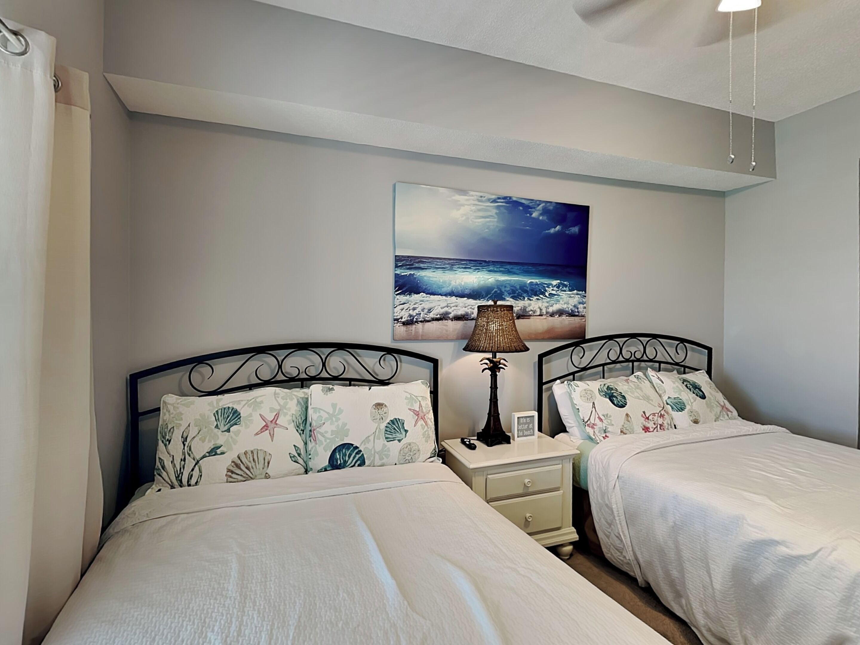 A bedroom with two beds, a nightstand, a lamp, and a beach-themed wall art.
