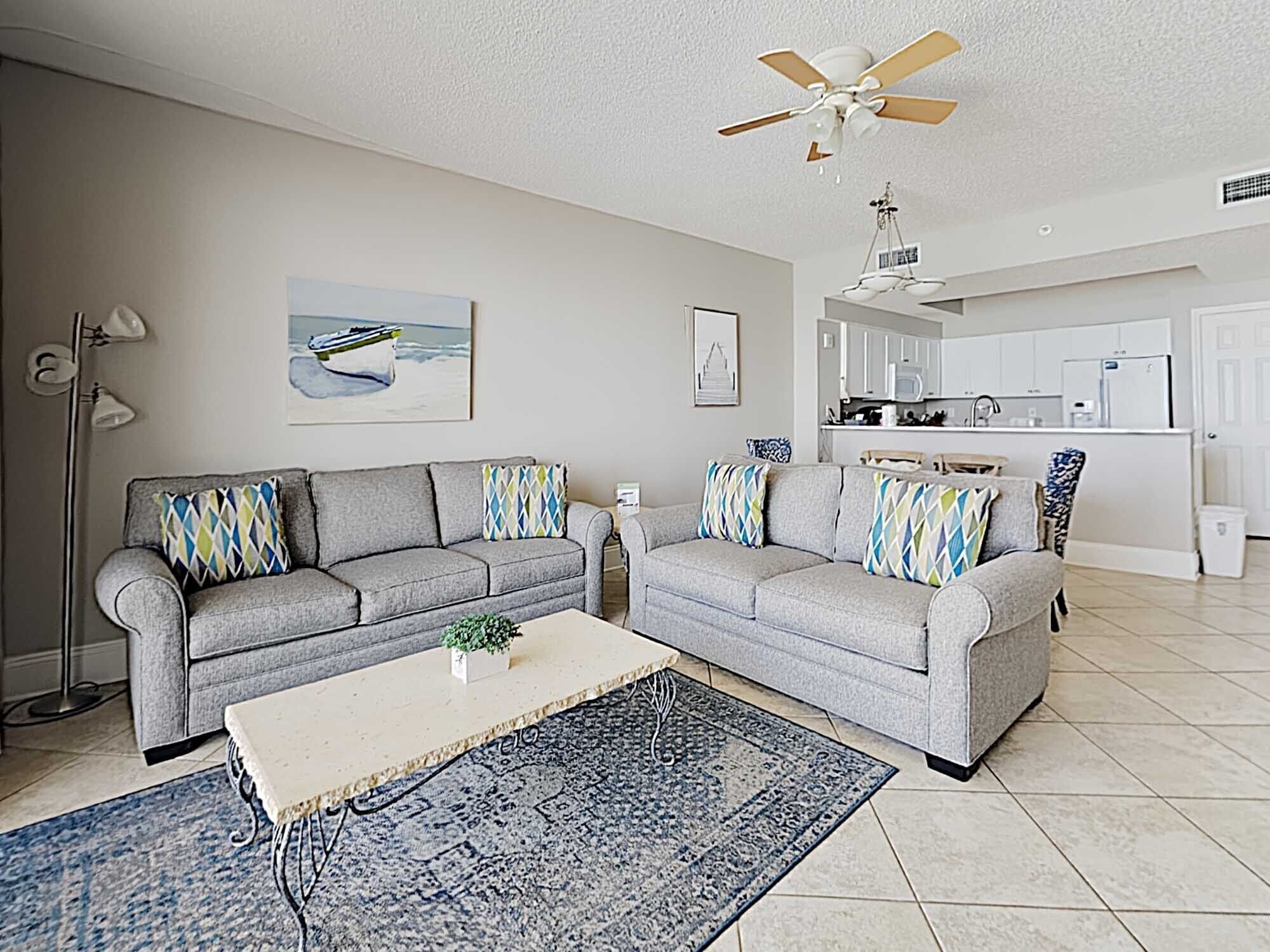 A living room with two grey sofas, a coffee table, a blue patterned rug, and a ceiling fan.