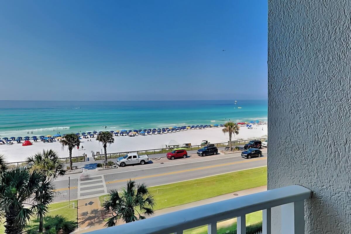 A beach view with a clear sky, a road with parked cars, and palm trees.