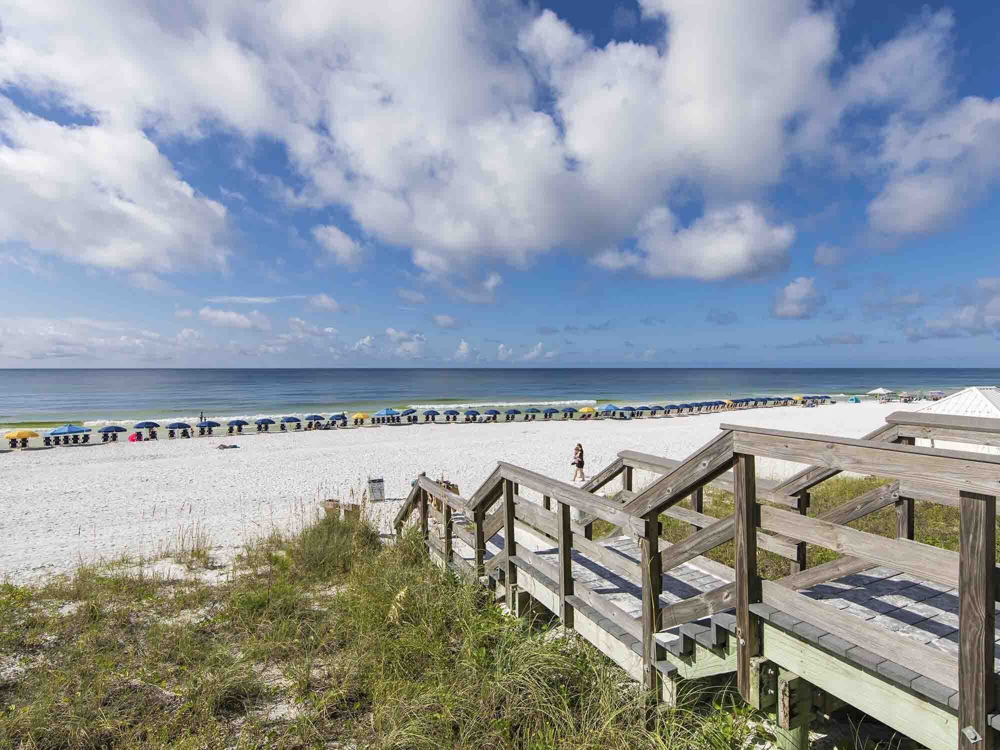 Une plage avec une promenade en bois menant au rivage, une rangée de parasols de plage et l’océan en arrière-plan.