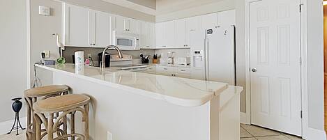 Una cocina moderna con armarios blancos, una isla central con taburetes, microondas, horno y refrigerador.