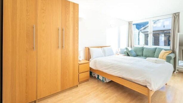 A bedroom with a wooden wardrobe, a bed with pillows, a bedside table, and a window with curtains. - GRACE SUITE LONDON (London)