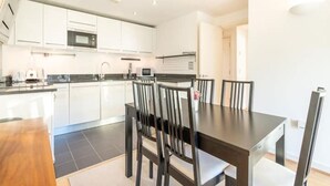 A modern kitchen with white cabinets, a central island, and a dining area with a dark wooden table and chairs. - GRACE SUITE LONDON (London)