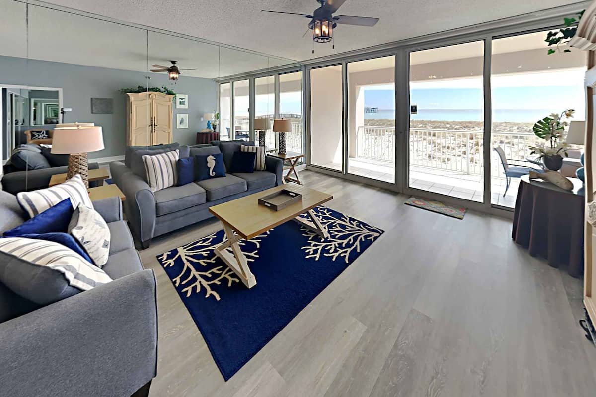 Un salon spacieux avec un canapé gris, une table basse et une vue sur la plage grâce à de grandes fenêtres.