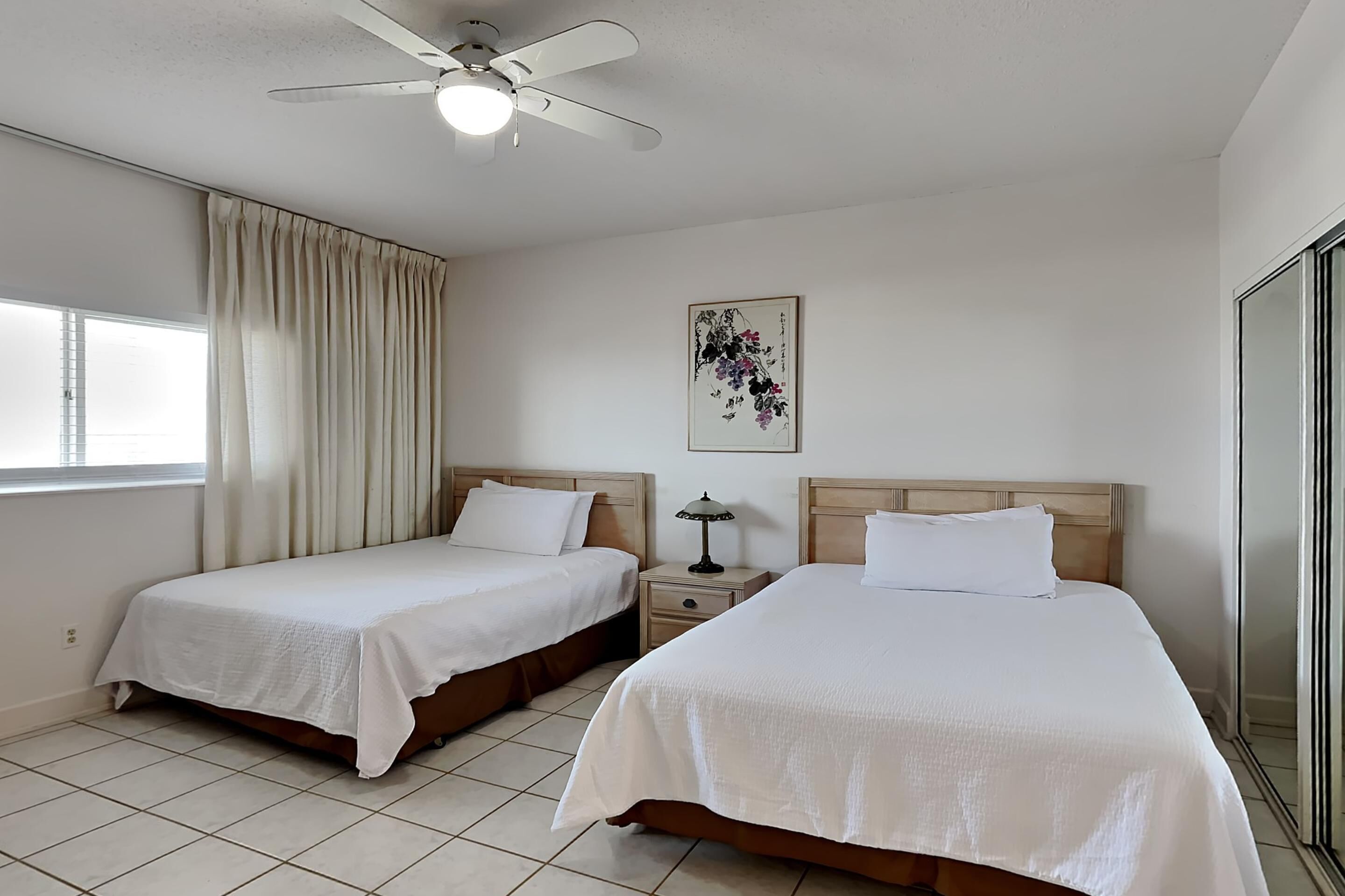 A hotel room with two beds, a ceiling fan, a window with curtains, a bedside table with a lamp, and a framed artwork on the wall.