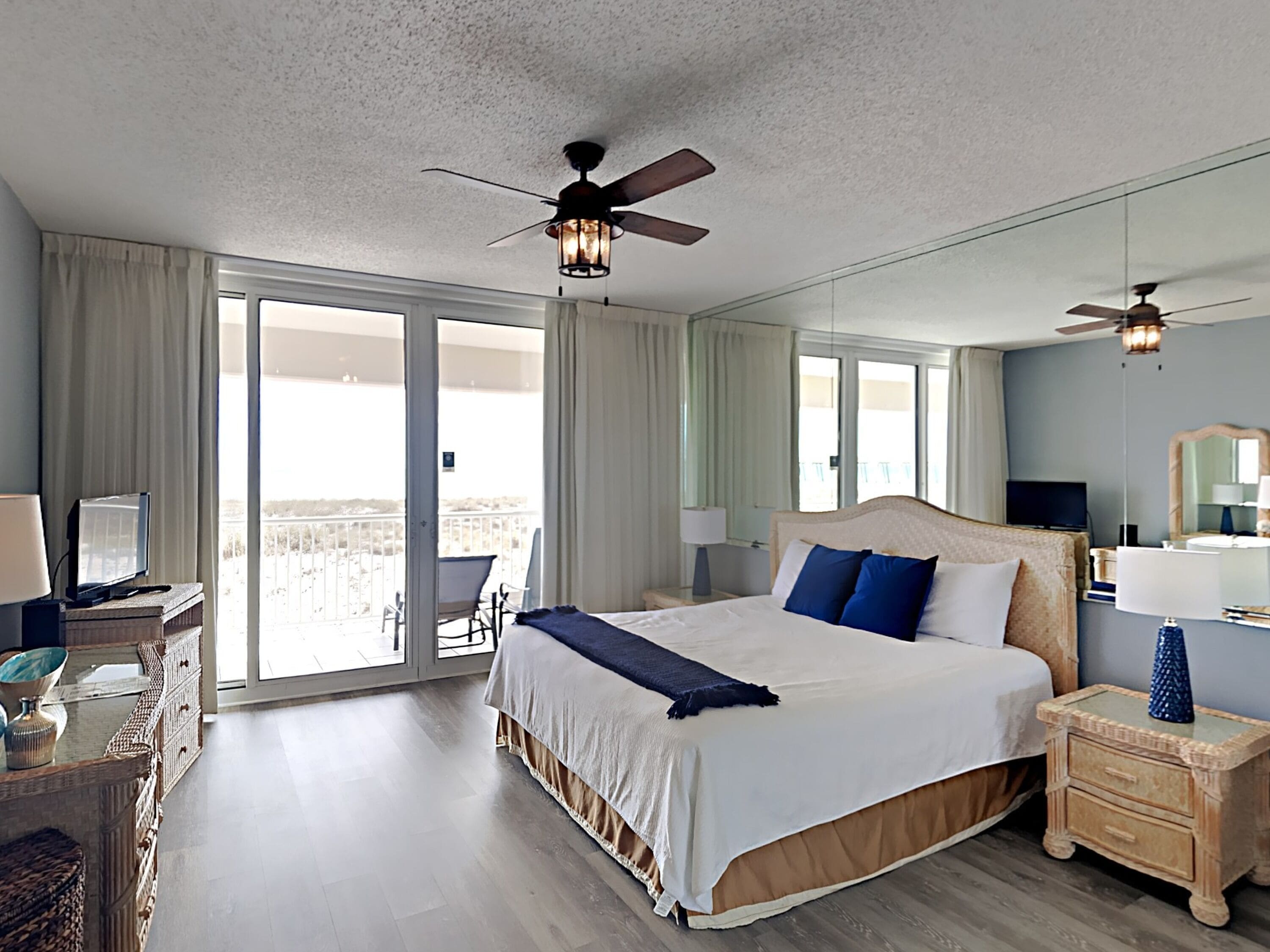 A spacious hotel room with a large bed, two ceiling fans, a balcony with a chair, and a television on a dresser.