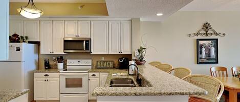 A kitchen with white cabinets, a microwave, an oven, a sink, and a dining area with wicker chairs.