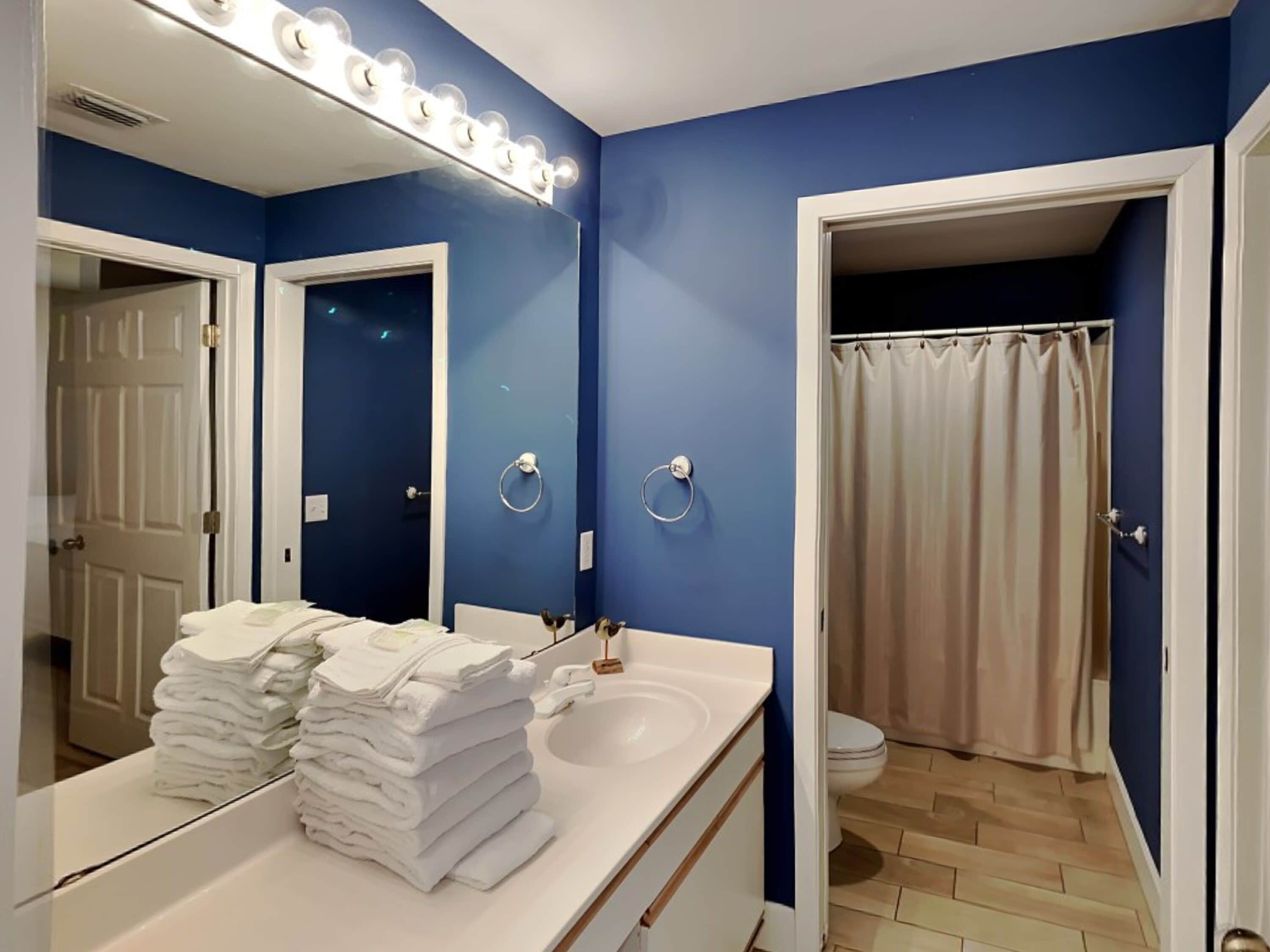 A bathroom with a blue wall, a white sink, a mirror with lights, a towel stack, and a shower curtain.