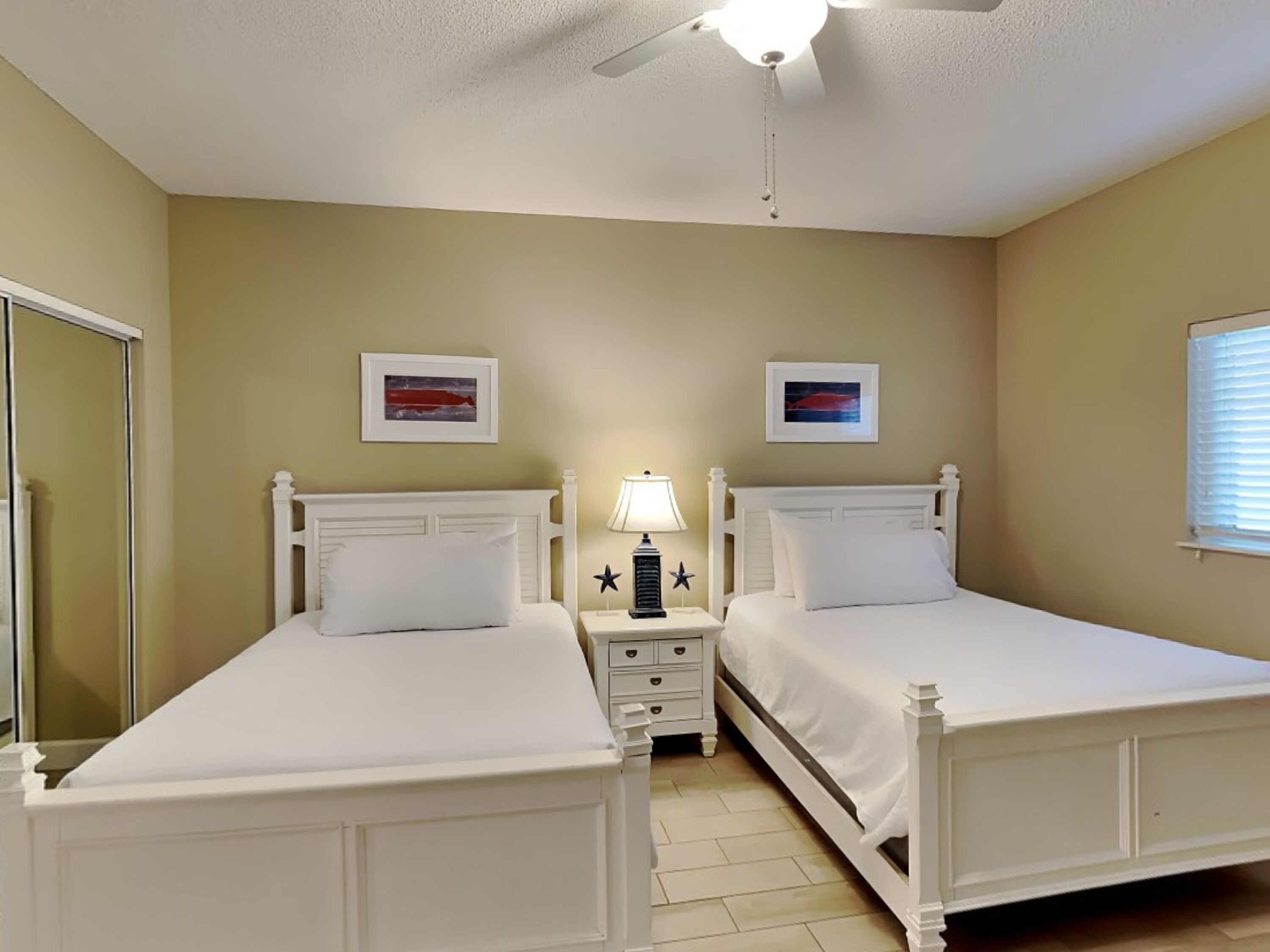 A twin bedroom with two beds, a nightstand with a lamp, and a ceiling fan.