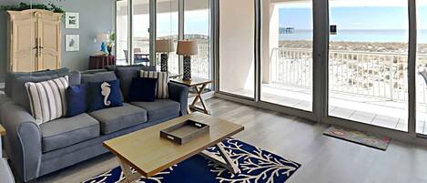 A spacious living room with a grey sofa, a wooden coffee table, and a large window offering a view of the beach.