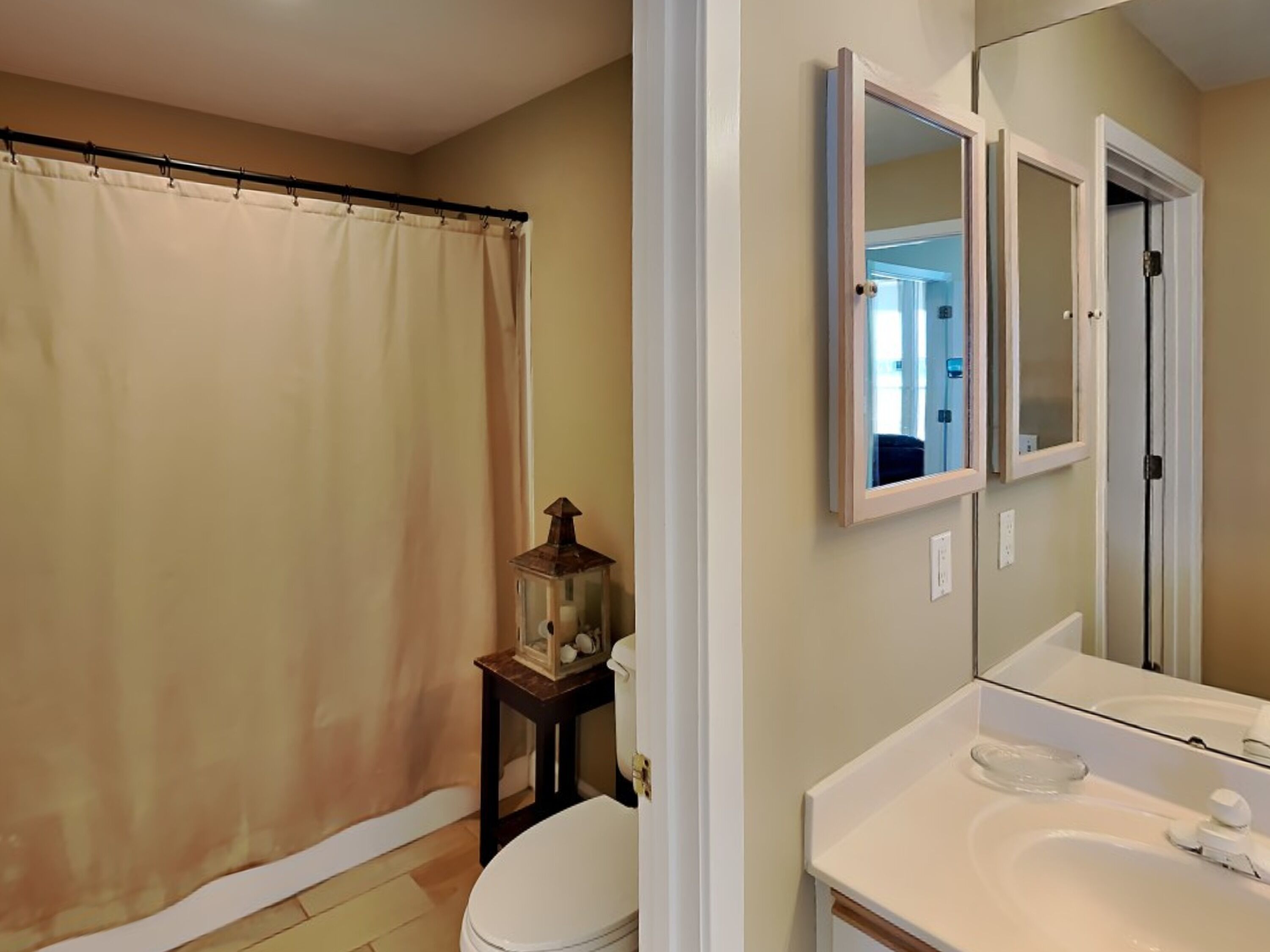 A bathroom with a shower curtain, a toilet, a small table with a lantern, and a mirror with a cabinet above it.