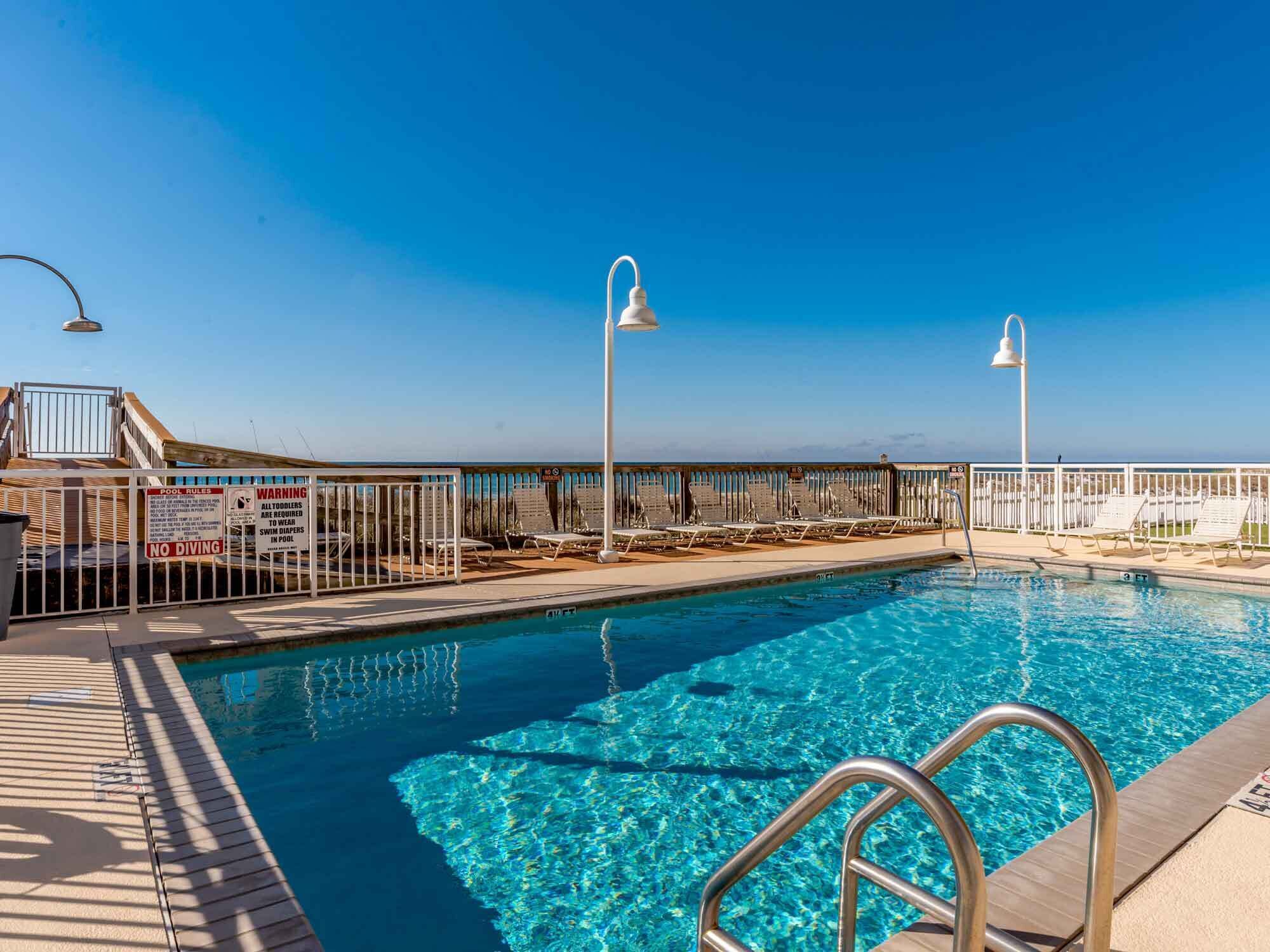 A clear blue pool with a lifeguard stand, lounge chairs, and a poolside area with a railing.