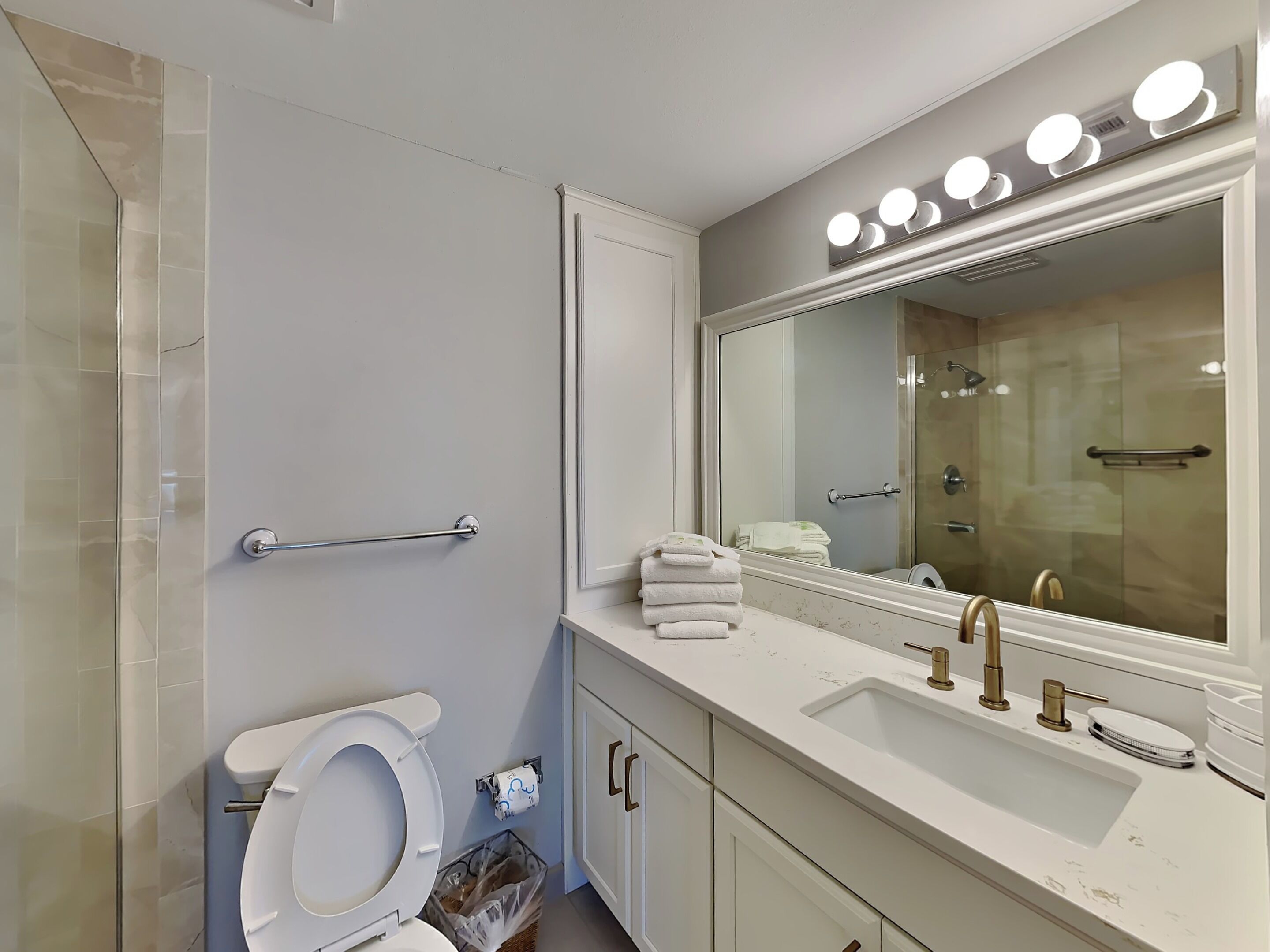 A modern bathroom with a white toilet, a sink with a gold faucet, and a vanity with a large mirror and lights.