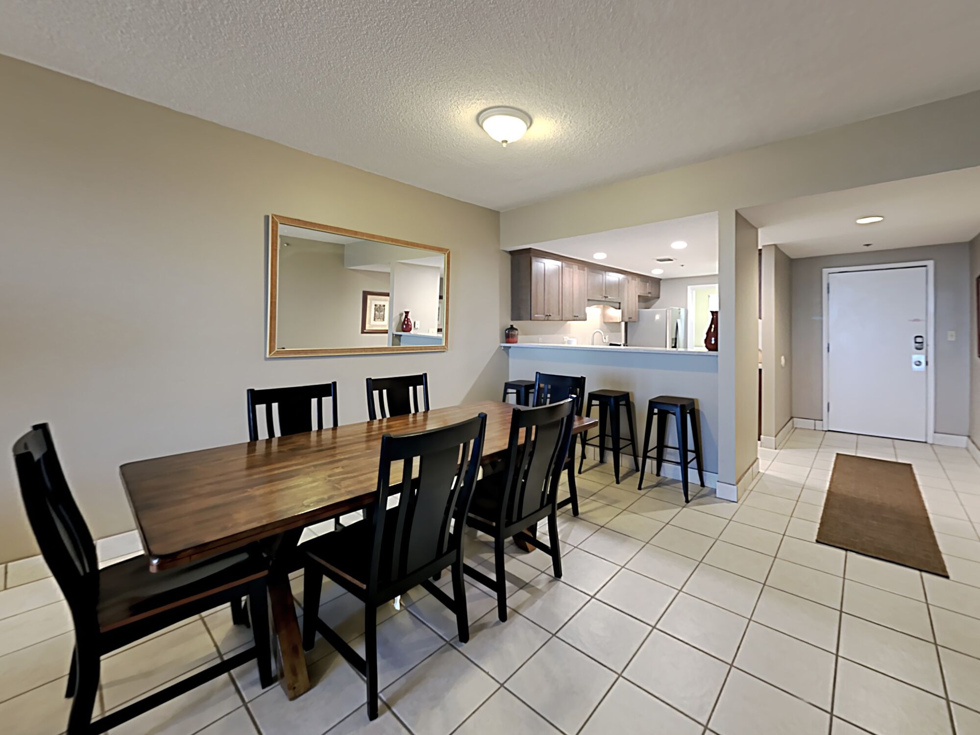 A dining area with a wooden table and chairs, a kitchen in the background, and a door leading to another room.